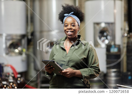 Portrait shot of positive female African American process technician wearing denim overall posing for camera while inspecting cider production taking notes on tablet computer at factory, copy space 120071658
