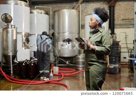 Side view of concentrated female African American process technician wearing denim overall inspecting tanks with craft cider while using tablet computer in workshop of factory, copy space Side view of concentrated female African American process technician wearing denim overall inspecting tanks with craft cider while using tablet computer in workshop of factory, copy space 120071665