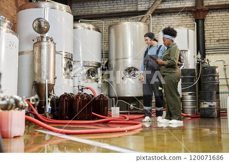 Wide angle shot of female African American process supervisor and male factory worker wearing denim overalls inspecting filling kegs with cider in workshop at plant 120071686