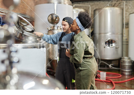 Medium shot of male factory worker opening lid of tank for female African American process technician to observe cider fermentation during quality inspection at plant, copy space 120071730