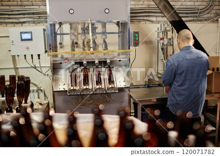 Rear view of male cider production operator directing bottling equipment filling beverage into bottles at factory 120071782