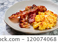 macro shot of fried bacon and scrambled eggs on a plate on a white marble serving table 120074068