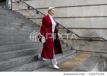Stylish Middle aged Woman in Burgundy Coat Walks City Street, Wearing Fashionable clothing White Turtleneck Sweater, Necklace and Grey Jeans, White Bag. Confident and Chic Mature Fashion Model Stylish Middle aged Woman in Burgundy Coat Walks City Street, Wearing Fashionable clothing White Turtleneck Sweater, Necklace and Grey Jeans, White Bag. Confident and Chic Mature Fashion Model 120075294
