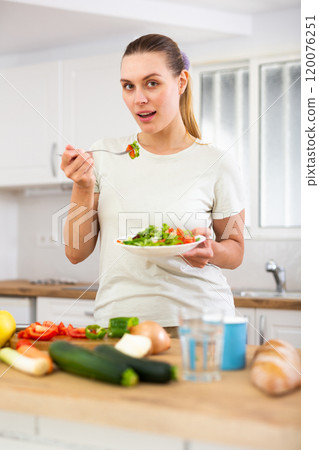 Positive young girl enjoying vegetable salad in home kitchen 120076251