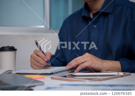 Businessman writing in notebook at office desk, planning and organizing ideas. Represents productivity, professional work environment. Ideal for corporate, educational uses. 120076385