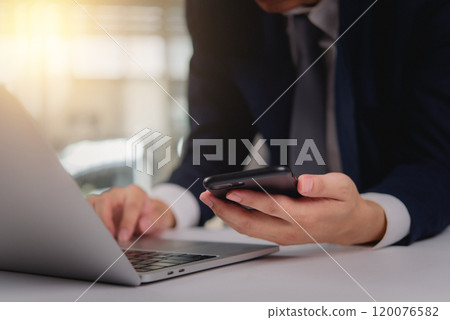 Business professional using a smartphone while working on a laptop. Suitable for modern business communication, remote work, or digital technology Business professional using a smartphone while working on a laptop. Suitable for modern business communication, remote work, or digital technology 120076582