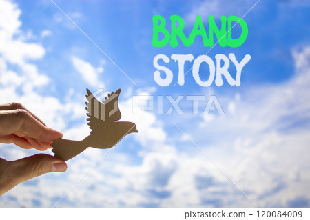 Brand story branding symbol. Concept words Brand story on beautiful blue sky. Beautiful blue sky clouds background. Hand holds wooden bird. Business brand story branding concept. Copy space. Brand story branding symbol. Concept words Brand story on beautiful blue sky. Beautiful blue sky clouds background. Hand holds wooden bird. Business brand story branding concept. Copy space. 120084009