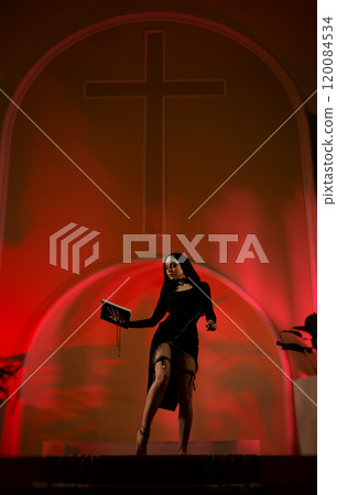 A nun girl in a sexy dress from an adult role-playing sex shop defiantly poses in a temple with a Bible book against the background of a cross 120084534