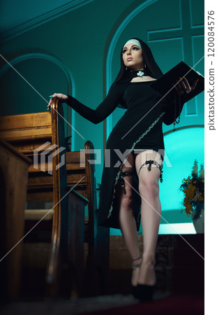 A nun in a sexy dress from an adult role-playing sex shop defiantly poses in a temple with a Bible book against the background of a cross 120084576
