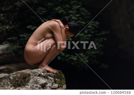 A beautiful naked Asian girl is squatting on a rock in the jungle with her arms wrapped around her legs 120084588