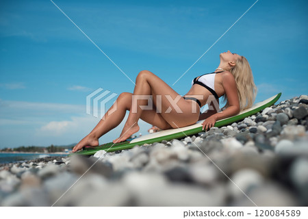 a beautiful girl in a swimsuit with a surfboard on the ocean shore against the background of a beautiful blue sky poses sexually 120084589