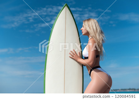 a beautiful girl in a swimsuit with a surfboard on the ocean shore against the background of a beautiful blue sky poses sexually 120084591