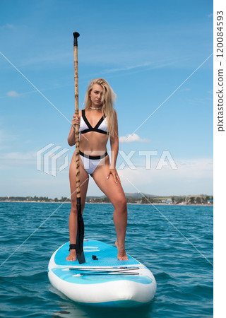 beautiful girl in a swimsuit with a sup board on the ocean shore against the background of a beautiful blue sky is rowing sexually with an oar beautiful girl in a swimsuit with a sup board on the ocean shore against the background of a beautiful blue sky is rowing sexually with an oar 120084593