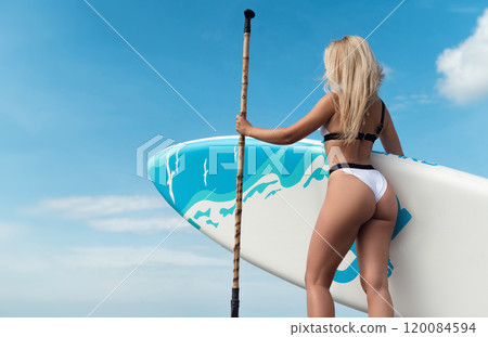 beautiful girl in a swimsuit with a sup board on the ocean shore against the background of a beautiful blue sky is rowing sexually with an oar beautiful girl in a swimsuit with a sup board on the ocean shore against the background of a beautiful blue sky is rowing sexually with an oar 120084594