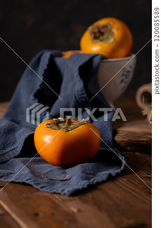 Ripe persimmon sits on a dark blue cloth, with a ceramic bowl in the background Ripe persimmon sits on a dark blue cloth, with a ceramic bowl in the background 120085589