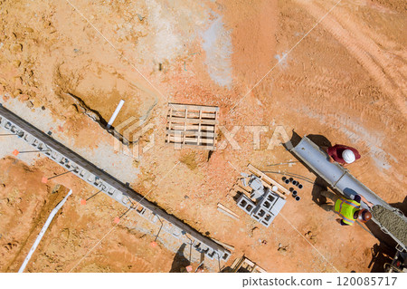 An working on construction uses a concrete mixer truck to pour concrete along foundational walls in area with reddish soil. An working on construction uses a concrete mixer truck to pour concrete along foundational walls in area with reddish soil. 120085717