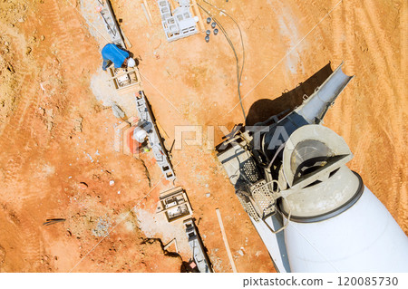 Worker uses mixer truck to pour concrete along foundation of building in large, reddish soil colored area. 120085730