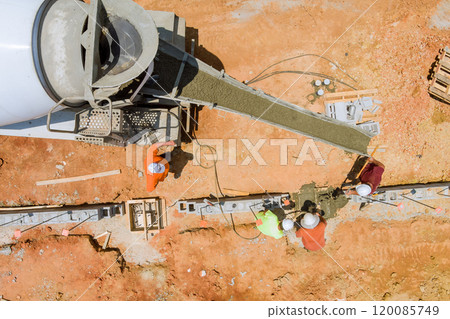 Construction worker pours concrete along foundation in an area with reddish soil using concrete mixer truck. 120085749