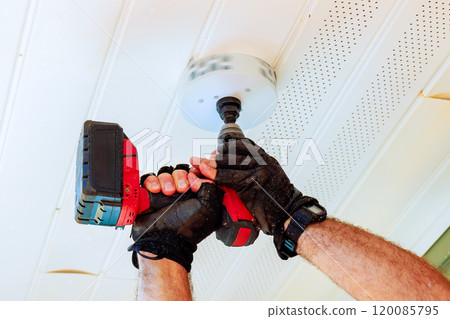 An individual wearing gloves holds cordless drill that has hole saw attachment attached, cuts circular hole in white ceiling to accommodate new light fixture 120085795