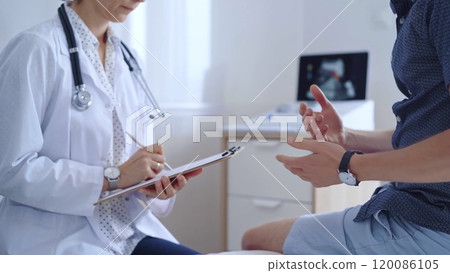Doctor woman is taking notes on a clipboard while a male patient describes his symptoms in clinic procedure cabinet. Medicine service 120086105