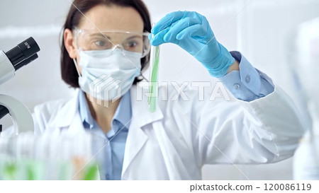 Woman scientist with protective gloves and glasses examining lab tube with a green fluid. Microbiology and science concept Woman scientist with protective gloves and glasses examining lab tube with a green fluid. Microbiology and science concept 120086119