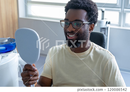 African American male patient looking at his teeth with dental mirror in modern dental office 120086542