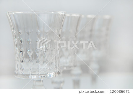 Crystal wine glasses with expanded edges and faceted pattern stand in a row on a white background, the first one is in focus on the sides, the second ones stand behind it in focus, close-up, the legs 120086671