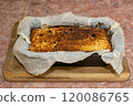 A baked cheesecake with a golden brown crust lies in a black pan on a wooden cutting board in a cozy kitchen setting. 120086765