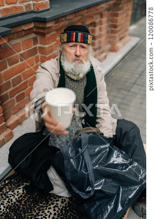 Vertical portrait of aged homeless man sits on urban street with belongings, seeking assistance, holding disposable cup and begging for alms. Concept of poor unhappy retirement. Vertical portrait of aged homeless man sits on urban street with belongings, seeking assistance, holding disposable cup and begging for alms. Concept of poor unhappy retirement. 120086778