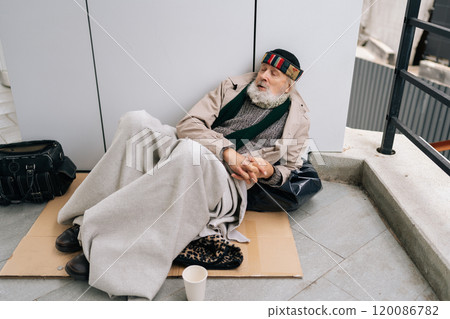 Tired elderly homeless man with long white beard sitting lying on sidewalk with belongings, holding cup and seeking assistance. Concept of poverty and vulnerability, evoking empathy and compassion. Tired elderly homeless man with long white beard sitting lying on sidewalk with belongings, holding cup and seeking assistance. Concept of poverty and vulnerability, evoking empathy and compassion. 120086782