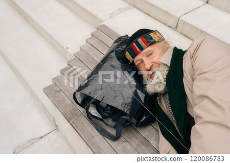 Top view of elderly homeless man sleeping on urban bench outside, using bag of belongings as pillow. Tired freezing aged male bundled up in coat and blanket, trying to stay warm, looking at camera 120086783