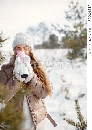 Young blonde woman standing on a nature at winter. Girl holding a pink thermos with a tea. Woman wearing brown jacket, grey hat and gloves. 120089411