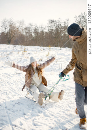 Portrait of romantic couple having fun on winter walk. Man carry his wife on a sled. Family wearing warm jacket and knitted hats. 120089447