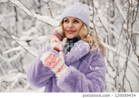 Portrait of young blonde woman standing at winter on a snow. Caucasian woman wearing purple warm fur coat and knitted hat. Woman posing for a photo. 120089454