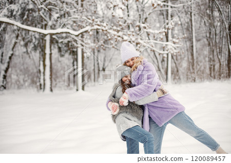 Young blonde woman with her daughter playing in winter in a snowballs. Caucasian woman wearing purple warm jacket and knitted hat. Brunette girl wearing grey jacket and hat. 120089457