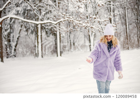 Portrait of young blonde woman standing at winter on a snow. Caucasian woman wearing purple warm fur coat and knitted hat. Woman posing for a photo. 120089458
