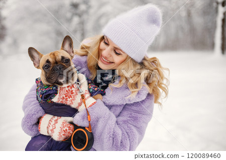 Young blonde woman with her dog french bulldog playing in winter on a snow. Caucasian woman wearing purple warm jacket and knitted hat. Dog wearing warm overalls. Young blonde woman with her dog french bulldog playing in winter on a snow. Caucasian woman wearing purple warm jacket and knitted hat. Dog wearing warm overalls. 120089460