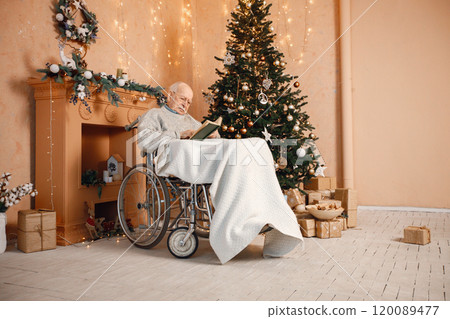 Old grey man celebrating Christmas alone. Man on a wheelchair sitting near Christmas tree. Grandfather covering on blanket and reading a book. 120089477