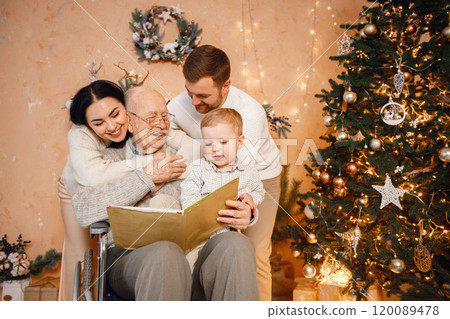Young family celebrating Christmas together. Brunette mother, bearded father, blonde son and old grandfather on a wheelchair sitting near Christmas tree and reading a book. Family wearing beige Young family celebrating Christmas together. Brunette mother, bearded father, blonde son and old grandfather on a wheelchair sitting near Christmas tree and reading a book. Family wearing beige 120089478