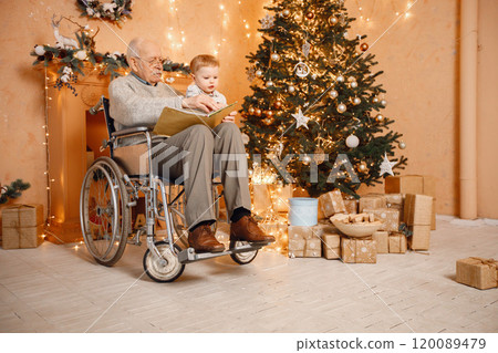 Little boy and his grandfather celebrating Christmas together. Blonde grandson and old grandfather on a wheelchair sitting near Christmas tree. Boy and grandfather reading a book. Little boy and his grandfather celebrating Christmas together. Blonde grandson and old grandfather on a wheelchair sitting near Christmas tree. Boy and grandfather reading a book. 120089479