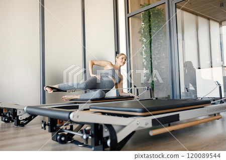 Pregnant woman doing exercises with a special equipment. Fit blonde woman working out in a gym. Woman wearing grey sportive leggins and top. 120089544