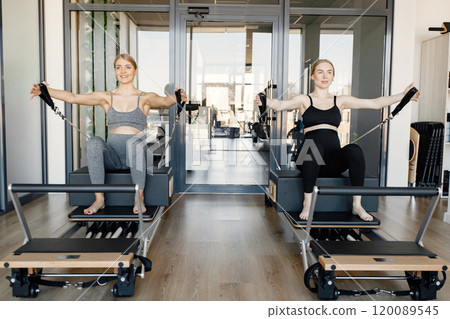 Two young pregnant women doing exercises on a special equipment. Fit blonde and brunette woman working out in a gym. Women wearing grey and black sportive leggins and tops. Two young pregnant women doing exercises on a special equipment. Fit blonde and brunette woman working out in a gym. Women wearing grey and black sportive leggins and tops. 120089545