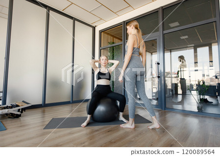 Two pregnant women doing stretching exercise. Fit blonde female coach helping with an excercises. Women wearing grey and black sportive leggins and tops. Two pregnant women doing stretching exercise. Fit blonde female coach helping with an excercises. Women wearing grey and black sportive leggins and tops. 120089564