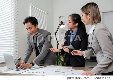 Three people in business attire are standing around a table with a laptop 120095153