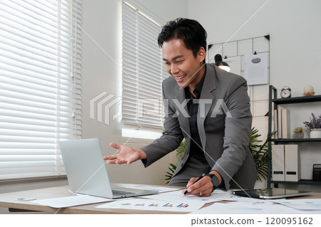 A man in a suit is sitting at a desk with a laptop open and a smile on his face 120095162