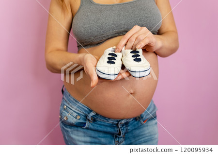 A joyful expectant mother holding tiny baby sneakers against her pregnant belly 120095934
