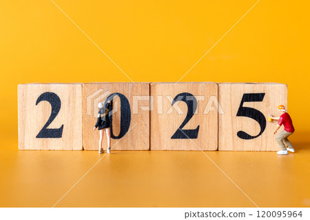 Miniature figures in overalls paint and construct the numbers 2025 on wooden blocks 120095964