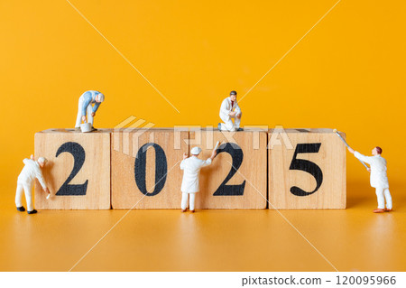 Miniature figures in overalls paint and construct the numbers 2025 on wooden blocks 120095966
