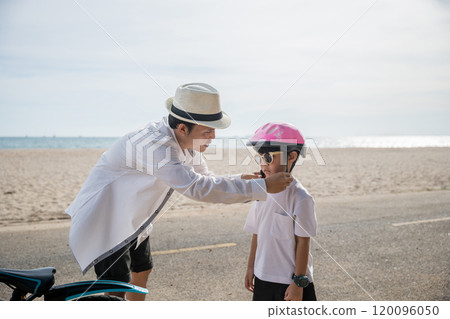 In the warm sunlight of beach day father dons a safety helmet offering inspiration and safety he teaches his cheerful son exciting skill of riding a bicycle a beautiful moment of family connection. 120096050