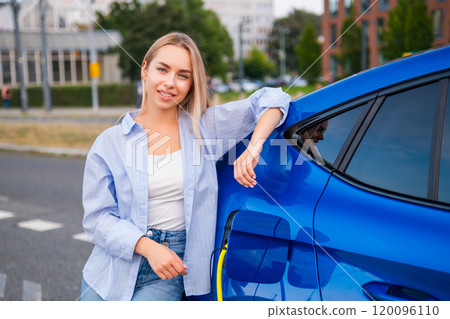 Blonde woman smiles while relaxing beside her electric vehicle as it charges in the street Blonde woman smiles while relaxing beside her electric vehicle as it charges in the street 120096110